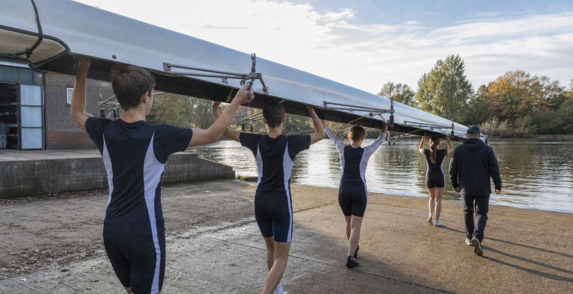 Young rowers carrying a racing shell beside a calm river